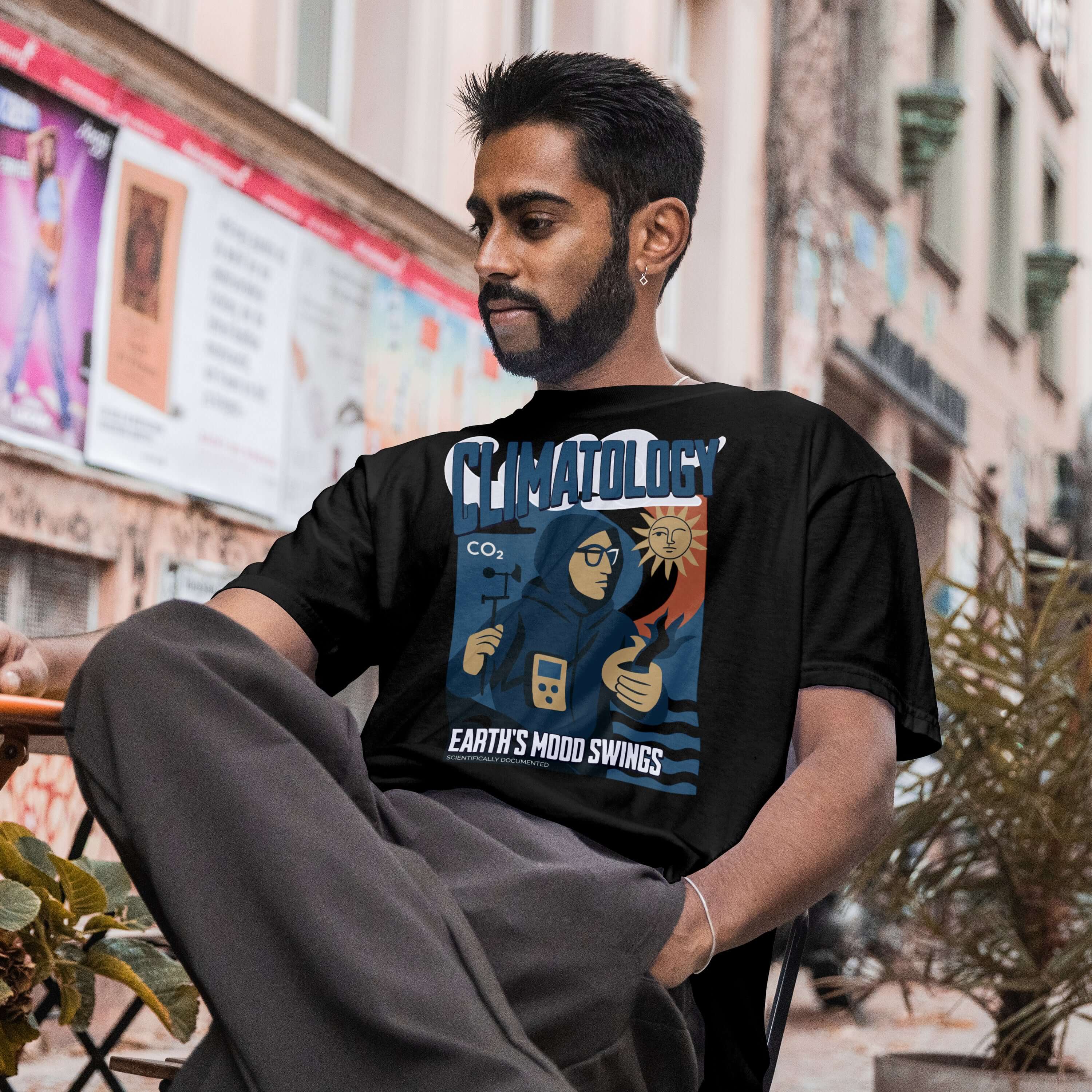 Man wearing Climatology T-Shirt with Earth's Mood Swings design, sitting outdoors. Celebrates climate scientists and their work.