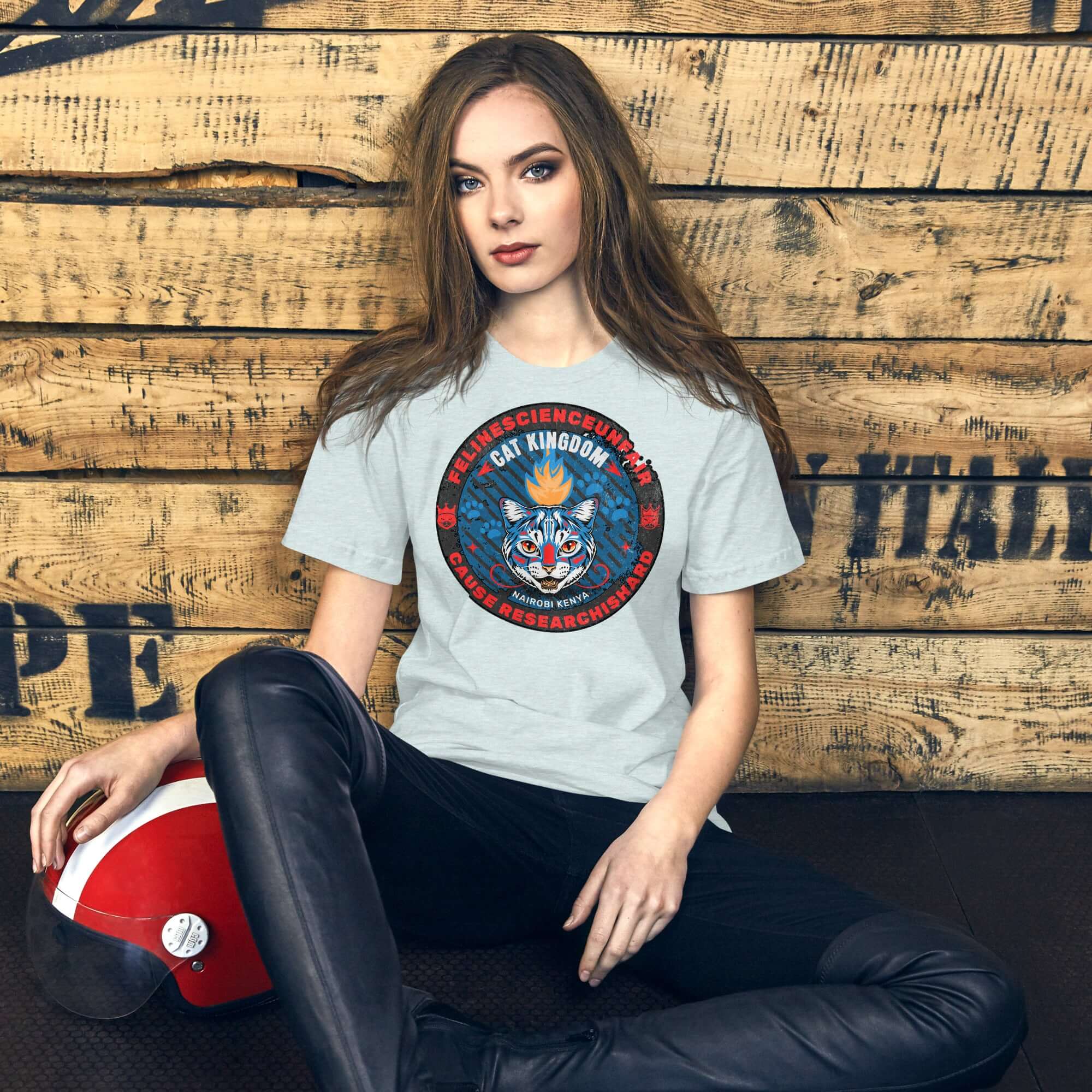 Woman wearing Feline Science Unit Cat Kingdom T-Shirt, featuring wildcat design, sitting against wooden wall in casual setting.
