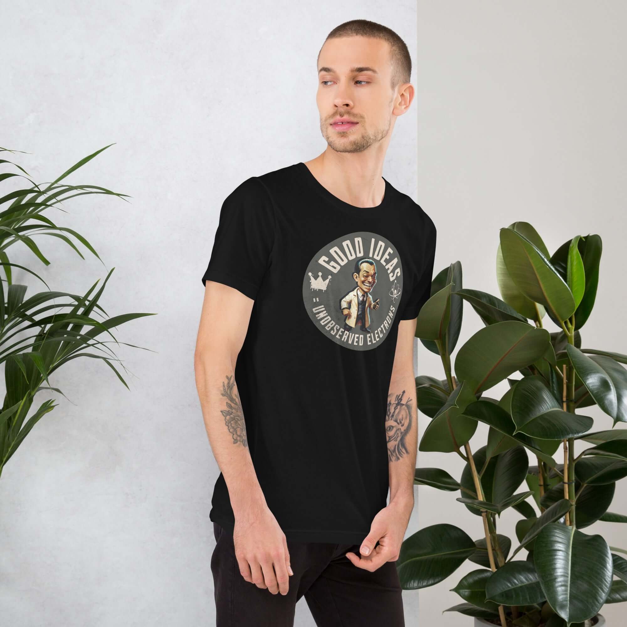 Man wearing "Good Ideas Unobserved Electrons" t-shirt with quantum physics humor, standing next to indoor plants.