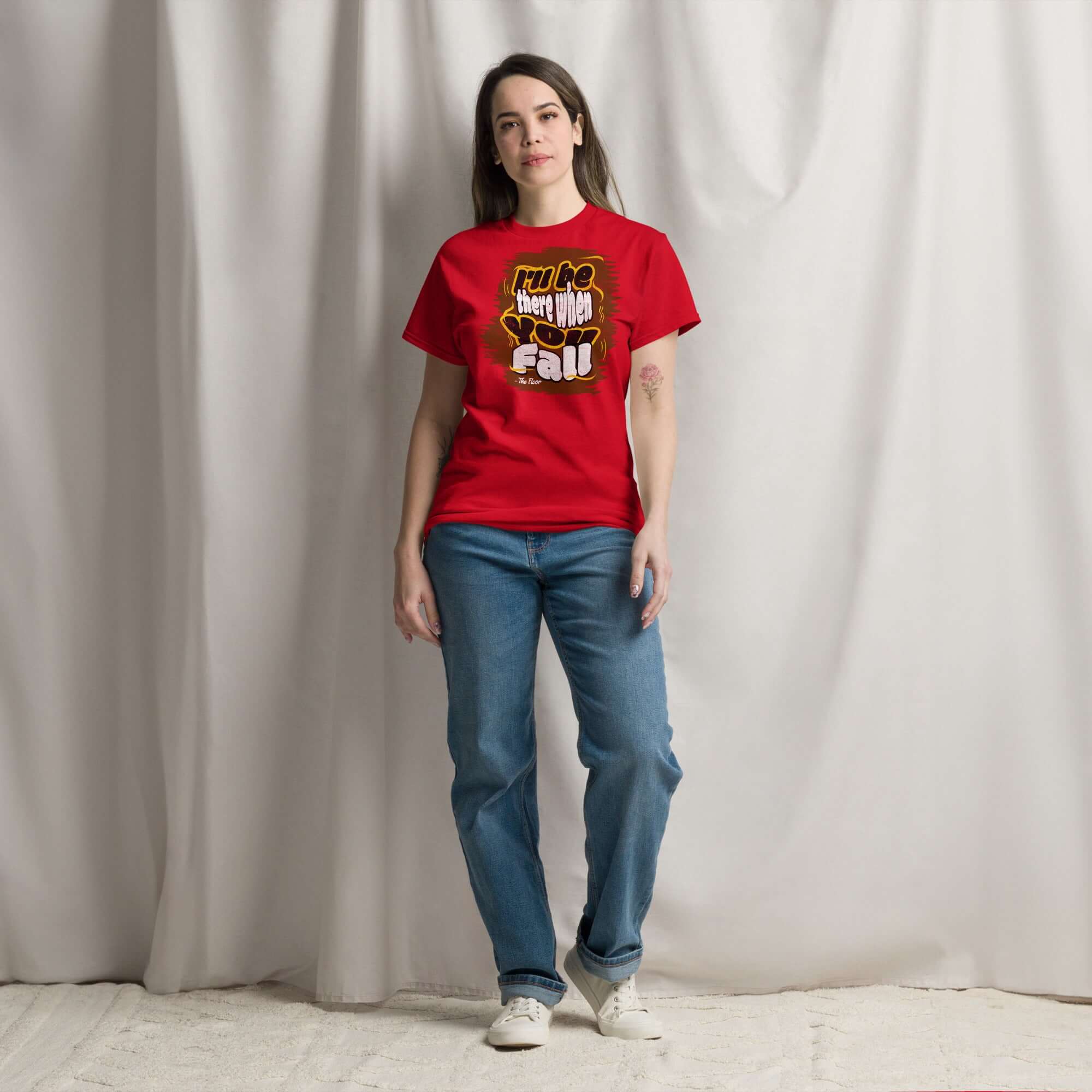 Woman wearing "I'll Be There When You Fall Floor" t-shirt, red Ironic Apparel, casual jeans, standing against white fabric backdrop.